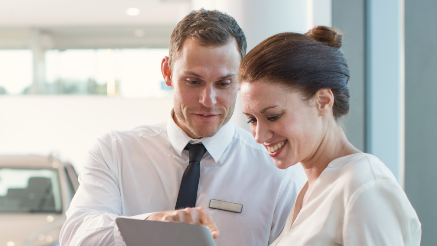 A man and woman are seen in a car showroom, inspecting a tablet.