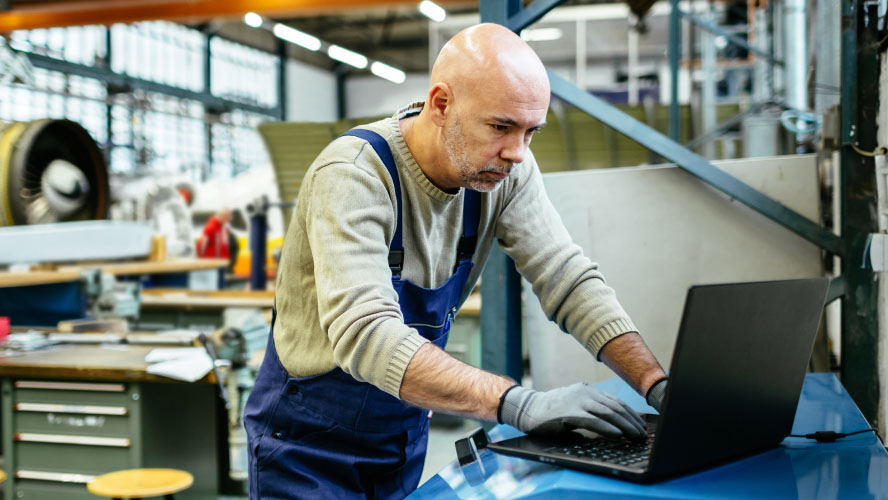 A man in an industrial setting focused on his laptop, diligently working on tasks.