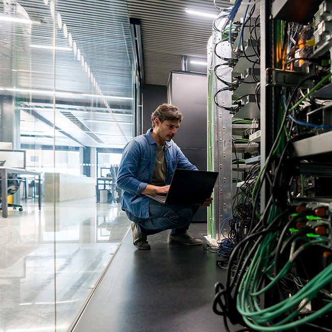 IT professional kneeling in front of a computer in a server room adjacent to an open office space