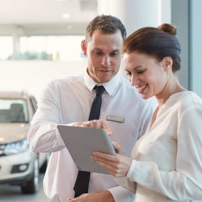 A man and woman are seen in a car showroom, inspecting a tablet.