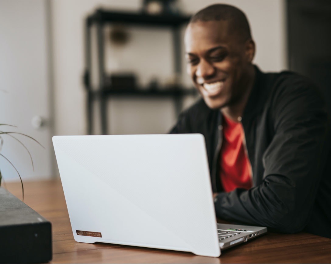 A man with a cheerful expression sits at a table, engrossed in his work on a laptop.