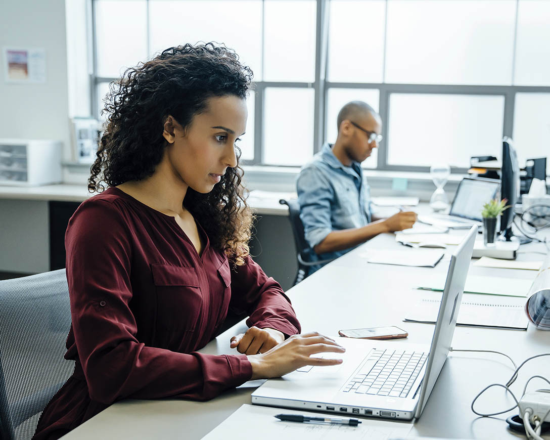 Recoverability – A woman works intently next to a colleague at a long desk in a bright office